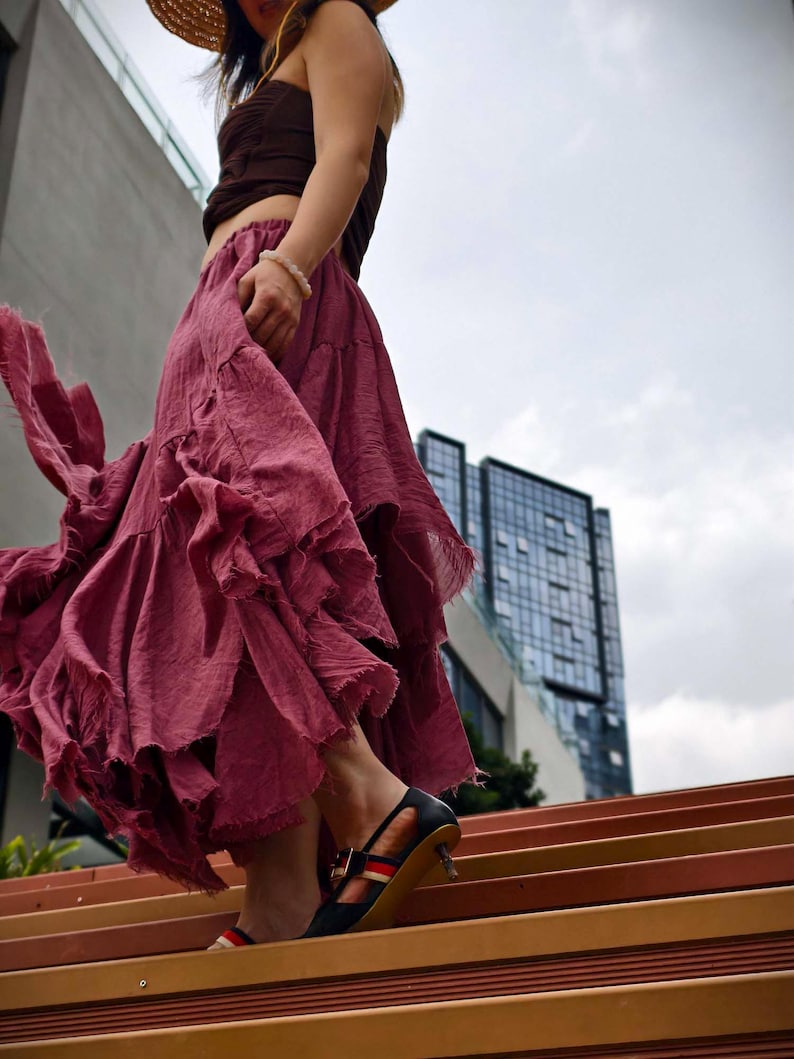 May include: A flowing, ruffled skirt in a deep rose color is paired with a brown top. The outfit is complemented by black heels with red and white stripes. The scene is set outdoors with a modern building in the background.