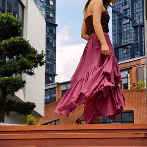 May include: A woman wearing a straw hat, brown tube top, and a flowing, ruffled, magenta skirt walks down wooden steps. The background includes modern buildings and a blue sky.