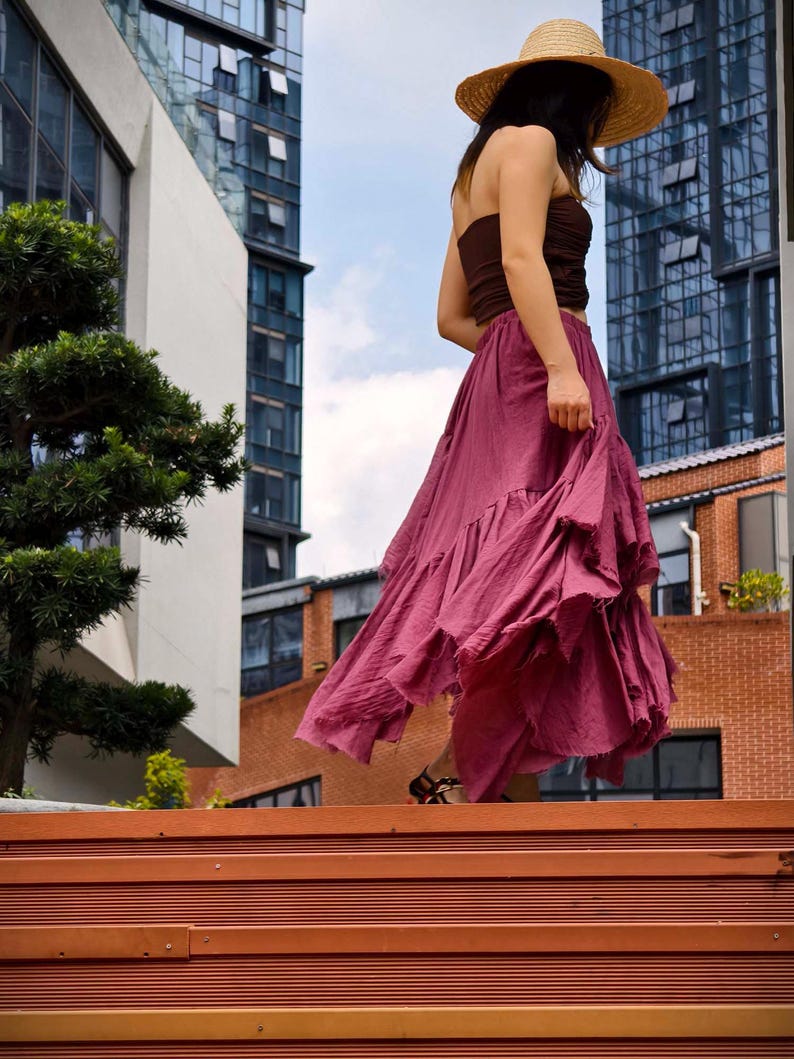 May include: A woman wearing a straw hat, brown tube top, and a flowing, ruffled, magenta skirt walks down wooden steps. The background includes modern buildings and a blue sky.