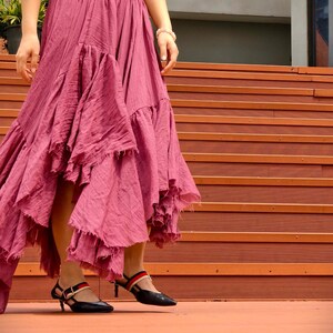 May include: A flowing, dusty rose skirt with multiple ruffled tiers. The skirt is paired with black heels featuring red and navy straps. The outfit is worn with a brown top. The model is standing on a wooden staircase.