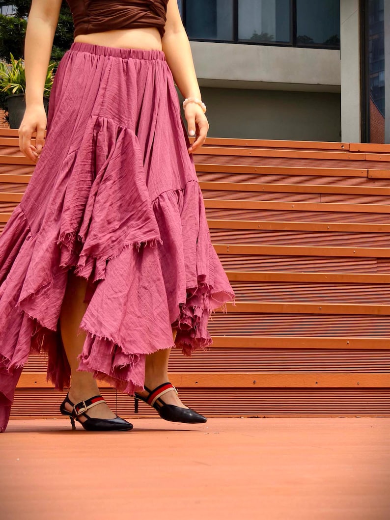 May include: A flowing, dusty rose skirt with multiple ruffled tiers. The skirt is paired with black heels featuring red and navy straps. The outfit is worn with a brown top. The model is standing on a wooden staircase.