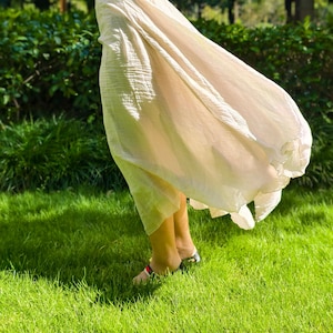 May include: A long, flowing, cream-colored skirt with a crinkled texture, paired with a black short-sleeved top. The skirt features a loose, voluminous silhouette that drapes down to the lower legs, creating a light and airy appearance in motion.