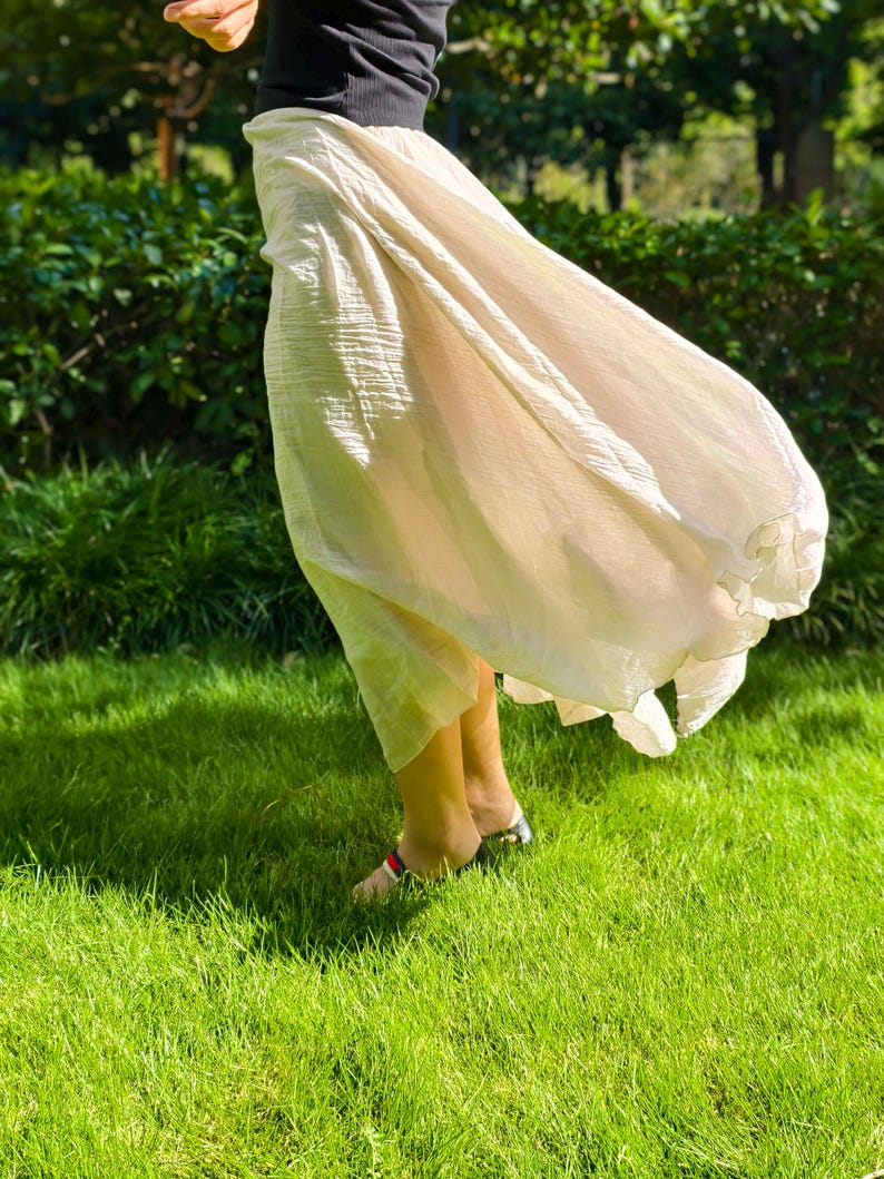 May include: A long, flowing, cream-colored skirt with a crinkled texture, paired with a black short-sleeved top. The skirt features a loose, voluminous silhouette that drapes down to the lower legs, creating a light and airy appearance in motion.