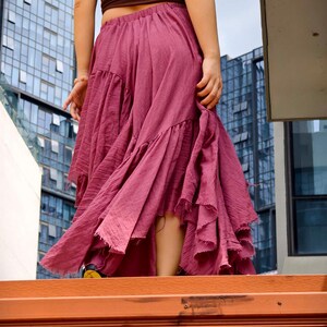 May include: A flowing, dusty rose skirt with multiple ruffled tiers. The skirt is paired with a brown tube top. The image is taken from a low angle, with a modern building in the background.