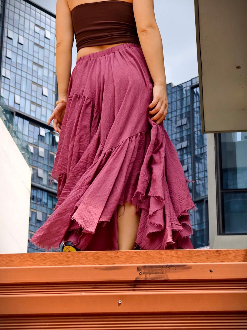 May include: A flowing, dusty rose skirt with multiple ruffled tiers. The skirt is paired with a brown tube top. The image is taken from a low angle, with a modern building in the background.