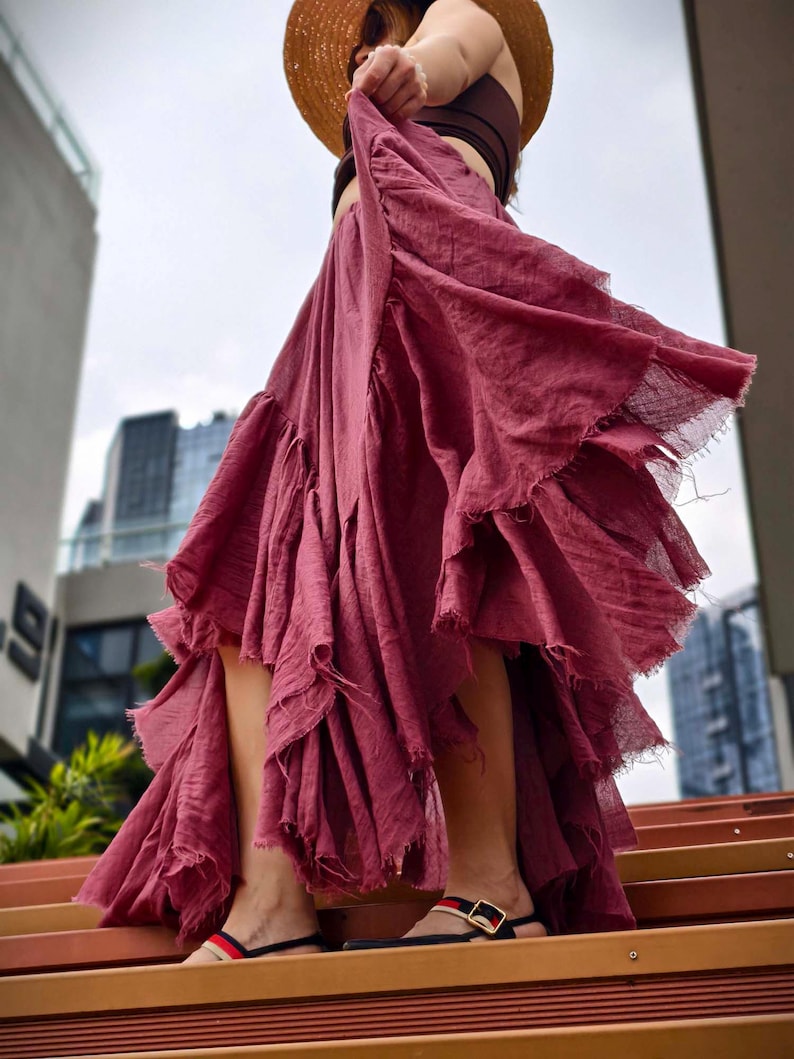 May include: A flowing, dusty rose-colored skirt with a tiered, ruffled design. The skirt has a frayed hemline and is paired with a brown top and straw hat. The image is taken from a low angle, with a city backdrop.
