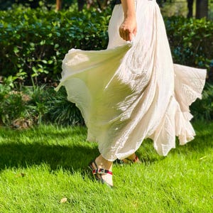 May include: A long, flowing, off-white skirt with a crinkled texture and a flared hemline. The skirt is paired with a black sleeveless top and black sandals featuring a red and green striped strap detail across the top of the foot.