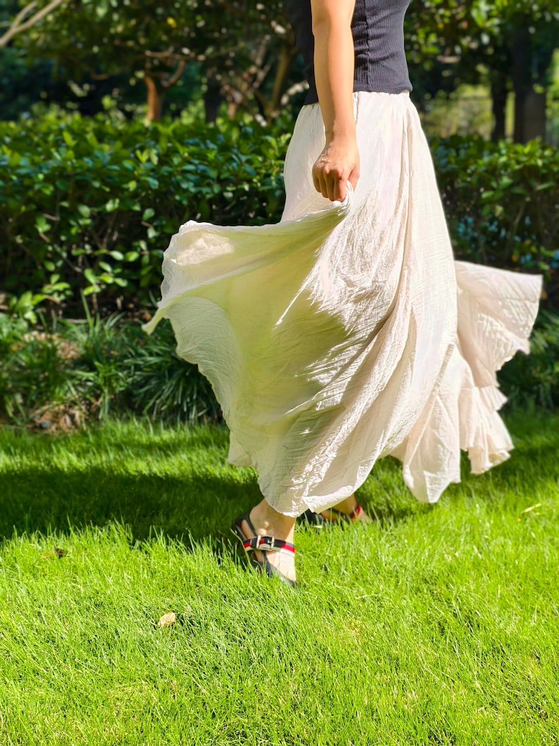 May include: A long, flowing, off-white skirt with a crinkled texture and a flared hemline. The skirt is paired with a black sleeveless top and black sandals featuring a red and green striped strap detail across the top of the foot.