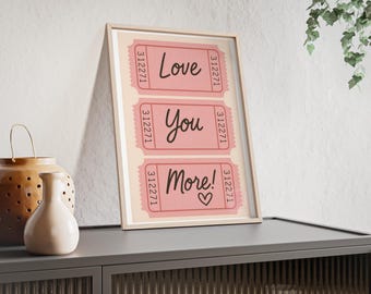 May include: Framed print featuring three pink ticket stubs with the words "Love You More!" in a handwritten style. The tickets have a light pink background and a white border. The print is in a light wood frame and sits on a gray cabinet.