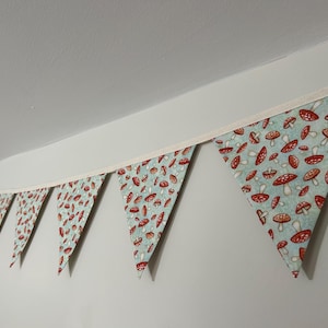 May include: A fabric bunting banner featuring a repeating pattern of red and white mushrooms on a light blue background. The triangular flags are attached to a cream-coloured fabric ribbon, creating a decorative string of pennants.