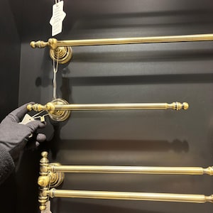Pode incluir: Exibi&ccedil;&atilde;o de toalheiros de lat&atilde;o antigo. Cada toalheiro apresenta uma barra horizontal com terminais decorativos e uma base circular montada na parede. Os toalheiros est&atilde;o dispostos verticalmente sobre um fundo escuro.