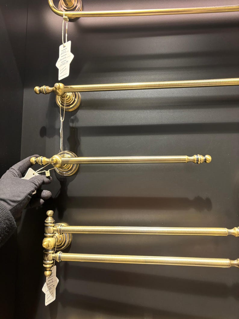 Pode incluir: Exibi&ccedil;&atilde;o de toalheiros de lat&atilde;o antigo. Cada toalheiro apresenta uma barra horizontal com terminais decorativos e uma base circular montada na parede. Os toalheiros est&atilde;o dispostos verticalmente sobre um fundo escuro.