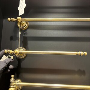 Pode incluir: V&aacute;rios toalheiros de lat&atilde;o antigo s&atilde;o montados numa parede escura. Cada toalheiro tem uma barra horizontal com terminais decorativos e placas de montagem circulares ornamentadas. Uma m&atilde;o com uma luva preta segura uma etiqueta.