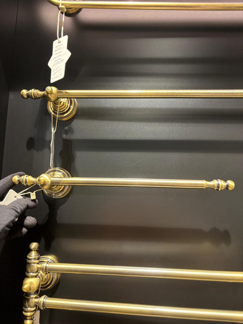 Pode incluir: V&aacute;rios toalheiros de lat&atilde;o antigo s&atilde;o montados numa parede escura. Cada toalheiro tem uma barra horizontal com terminais decorativos e placas de montagem circulares ornamentadas. Uma m&atilde;o com uma luva preta segura uma etiqueta.