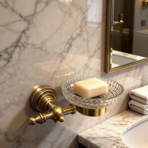 May include: A vintage-style soap dish with a decorative, ornate brass fixture. The clear, patterned glass dish holds a bar of soap. The fixture is mounted on a marble wall, with a mirror and sink visible in the background.