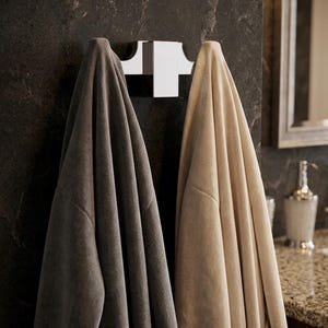 May include: A modern bathroom scene featuring a chrome towel rack mounted on a dark, textured wall. Two plush towels, one gray and one beige, hang from the rack. The scene includes a granite countertop, a mirror, and a soap dispenser.