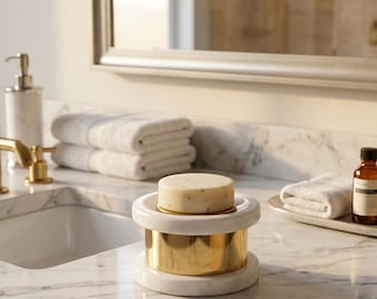 Può includere: Un portasapone in marmo e oro con una saponetta rotonda. Il portasapone ha una parte superiore e inferiore in marmo bianco con un cilindro in metallo dorato al centro. Il portasapone è su un piano di lavoro in marmo in un bagno.