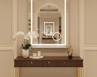 Könnte beinhalten: Ein rechteckiger, beleuchteter Spiegel mit weißem Rand hängt über einer Konsole aus dunklem Holz mit goldenen Beinen. Auf dem Tisch stehen eine weiße Vase mit Orchideen, Kerzen und eine dekorative goldene Skulptur. Der Spiegel spiegelt einen Flur wider.