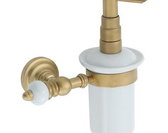 Puede incluir: Un dispensador de jabón montado en la pared con una botella de cerámica blanca y una bomba y herrajes dorados. El dispensador tiene un diseño clásico con un brazo ornamentado y un pomo de cerámica blanca.