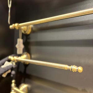 Pode incluir: Close-up de v&aacute;rias barras de toalha de lat&atilde;o antigo montadas em uma superf&iacute;cie escura. As barras t&ecirc;m um design cl&aacute;ssico com pe&ccedil;as decorativas nas extremidades e ferragens de montagem. O acabamento &eacute; dourado envelhecido.