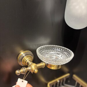 May include: Antique brass soap dish with a clear, textured glass bowl. The dish is mounted on a wall and features ornate detailing. A small tag is attached to the fixture.