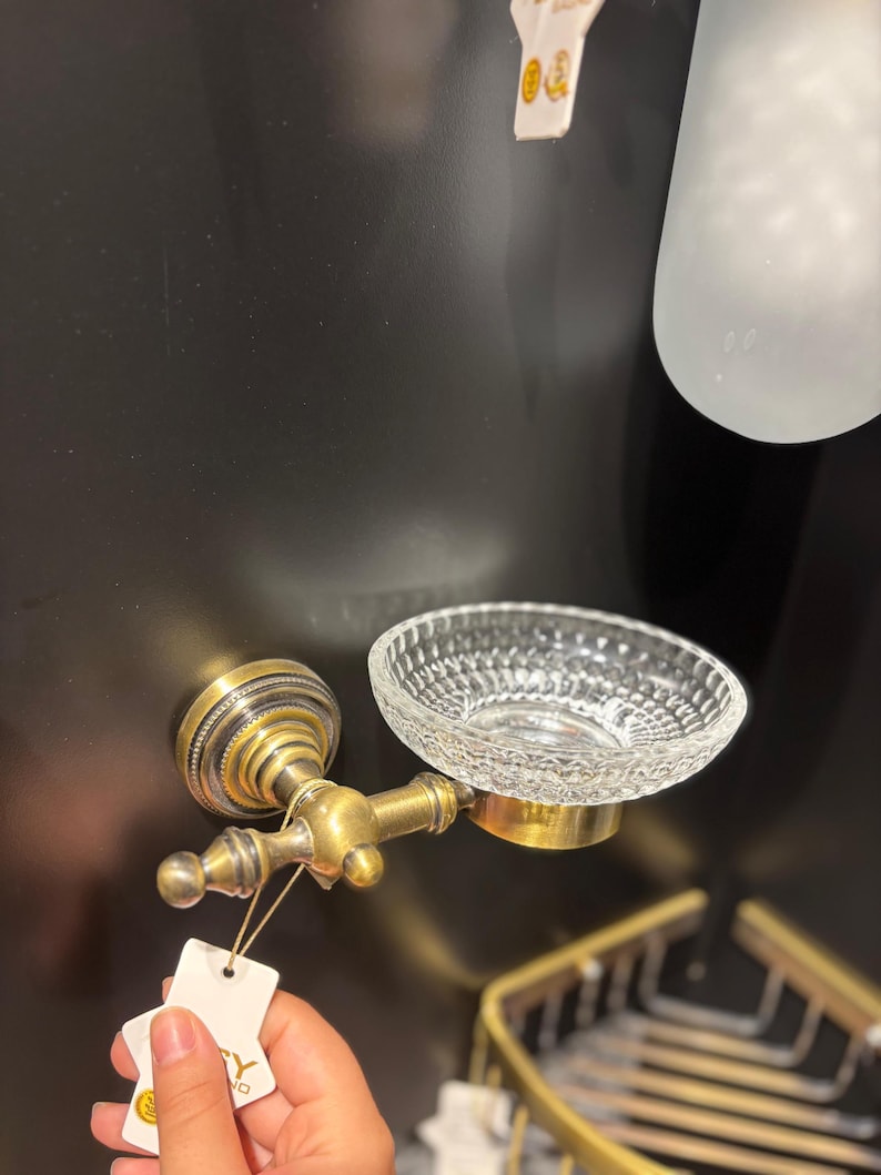 May include: Antique brass soap dish with a clear, textured glass bowl. The dish is mounted on a wall and features ornate detailing. A small tag is attached to the fixture.