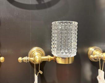 Puede incluir: Un vaso de vidrio transparente con un patrón texturizado se encuentra sobre un soporte de color latón. El soporte está montado en una pared oscura, y un anillo de latón a juego es visible en el fondo. El vaso está diseñado para uso en el baño.