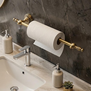 Pode incluir: Cena de casa de banho com um porta-rolo de papel higi&eacute;nico em lat&atilde;o e um rolo de papel branco. Uma pia branca, uma torneira cromada e dois dispensadores de sabonete texturizados. Um pequeno vaso dourado com uma planta suculenta.