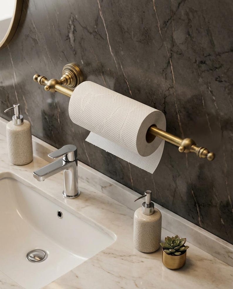 Pode incluir: Cena de casa de banho com um porta-rolo de papel higi&eacute;nico em lat&atilde;o e um rolo de papel branco. Uma pia branca, uma torneira cromada e dois dispensadores de sabonete texturizados. Um pequeno vaso dourado com uma planta suculenta.