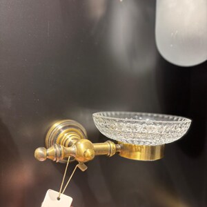 May include: A vintage-style soap dish with a clear, textured glass bowl and a brass-colored metal frame. The dish is mounted on a wall and features ornate detailing, suggesting a classic bathroom fixture.