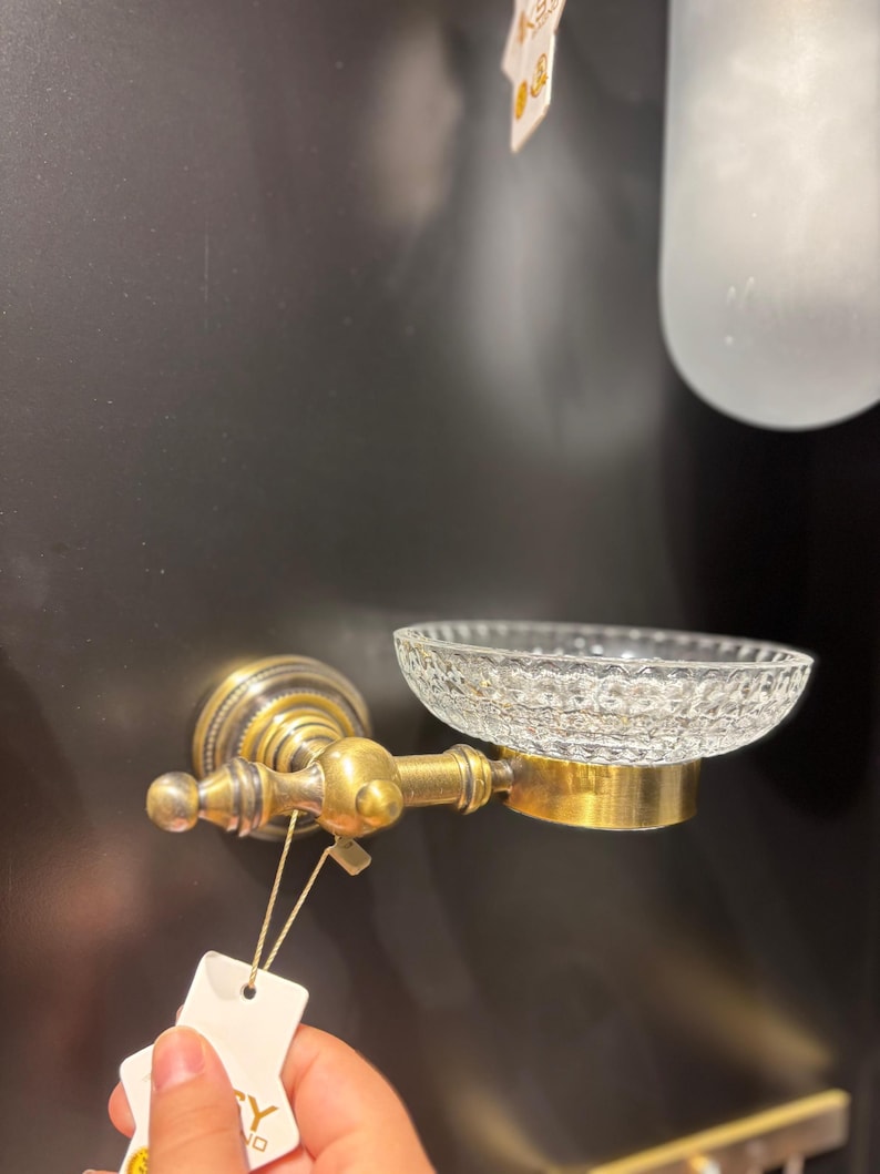 May include: A vintage-style soap dish with a clear, textured glass bowl and a brass-colored metal frame. The dish is mounted on a wall and features ornate detailing, suggesting a classic bathroom fixture.
