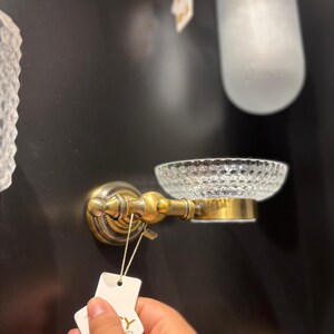 May include: A vintage-style soap dish with a clear, textured glass bowl and ornate brass hardware. The dish is mounted on a dark wall, showcasing its classic design. A tag is attached to the soap dish.