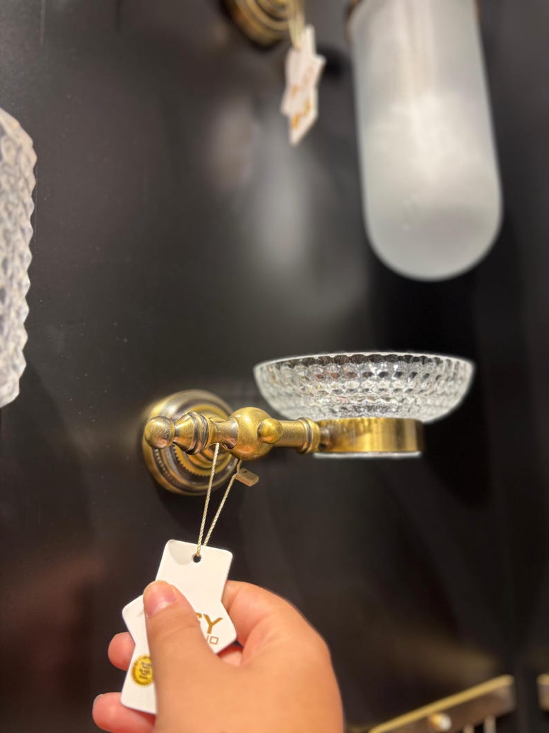May include: A vintage-style soap dish with a clear, textured glass bowl and ornate brass hardware. The dish is mounted on a dark wall, showcasing its classic design. A tag is attached to the soap dish.