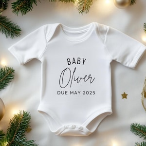 May include: White baby onesie with the text "BABY Oliver DUE MAY 2025" in black script. The onesie is surrounded by Christmas decorations, including evergreen branches, gold ornaments, and string lights.