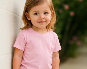 May include: A young child wearing a short-sleeved, light pink t-shirt and blue jeans. The child is smiling and leaning against a white wall. The background is blurred with greenery.