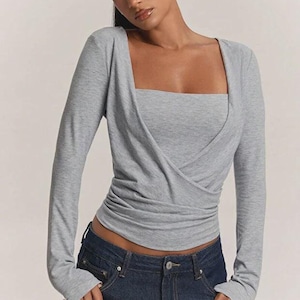 May include: A long-sleeved heather gray top featuring a square neckline and a wrap-style front with gathered fabric detailing at the waist. The garment is paired with dark blue denim jeans that have a visible metal button and zipper fly closure.