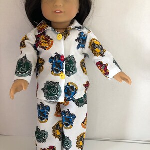 May include: A white pajama set for a doll, featuring a long-sleeved shirt and pants with a repeating pattern of the Hogwarts house crests. The crests are in their traditional colors of red, yellow, blue, and green.