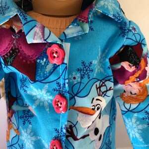 May include: A blue pajama top with a Frozen theme. The fabric features images of Elsa, Anna, and Olaf. The top has a button closure and pink buttons.