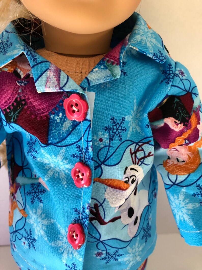 May include: A blue pajama top with a Frozen theme. The fabric features images of Elsa, Anna, and Olaf. The top has a button closure and pink buttons.