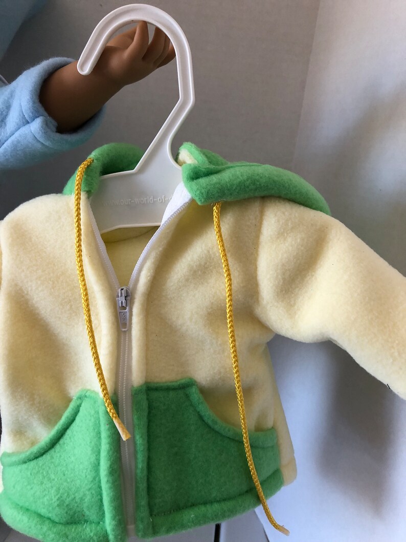 May include: A yellow and green fleece hoodie with a zipper closure and yellow drawstrings. The hoodie has a pocket on each side and the text "our-world-of" is visible on the zipper.