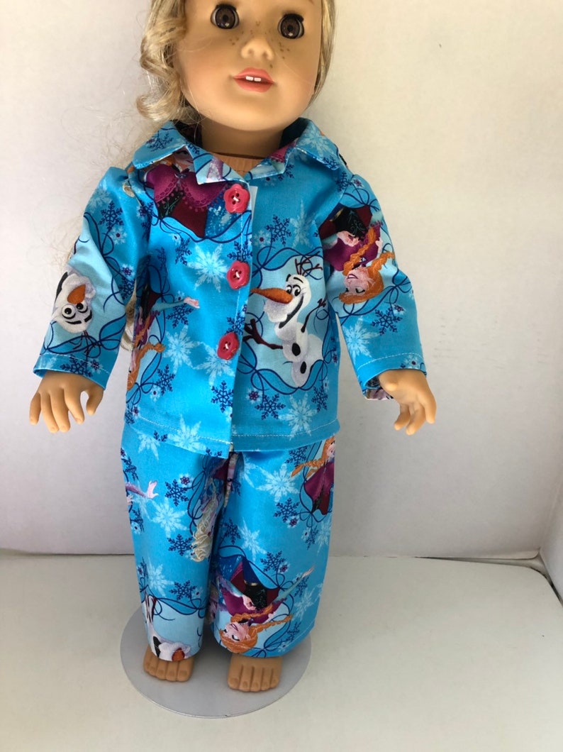 May include: A blue and white pajama set for a doll, featuring a snowflake pattern and images of Elsa, Anna, and Olaf from the movie Frozen.