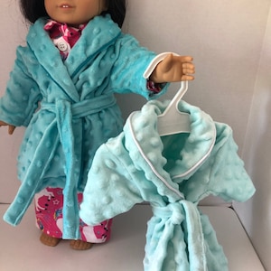 May include: A doll wearing a light blue plush robe with a matching belt and white trim. The robe has a textured, dotted pattern. A second robe on a hanger is also visible. The doll is wearing pajamas with a pink and white unicorn pattern.