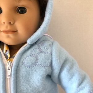 May include: A light blue hooded jacket with a white zipper and a stitched design on the front. The jacket has a pocket on the front.