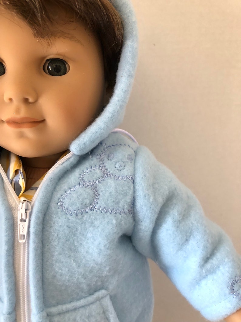 May include: A light blue hooded jacket with a white zipper and a stitched design on the front. The jacket has a pocket on the front.