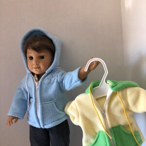 May include: Two fleece hooded jackets for dolls. One is light blue with a white zipper and the other is yellow, green, and white with a yellow zipper.