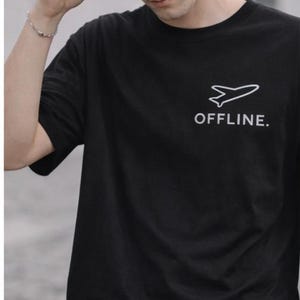 Puede incluir: Camiseta negra con un gr&aacute;fico de avi&oacute;n blanco y la palabra "OFFLINE". Se usa una gorra de b&eacute;isbol negra con el logotipo blanco de Nike. La imagen muestra a una persona usando la camiseta y la gorra.