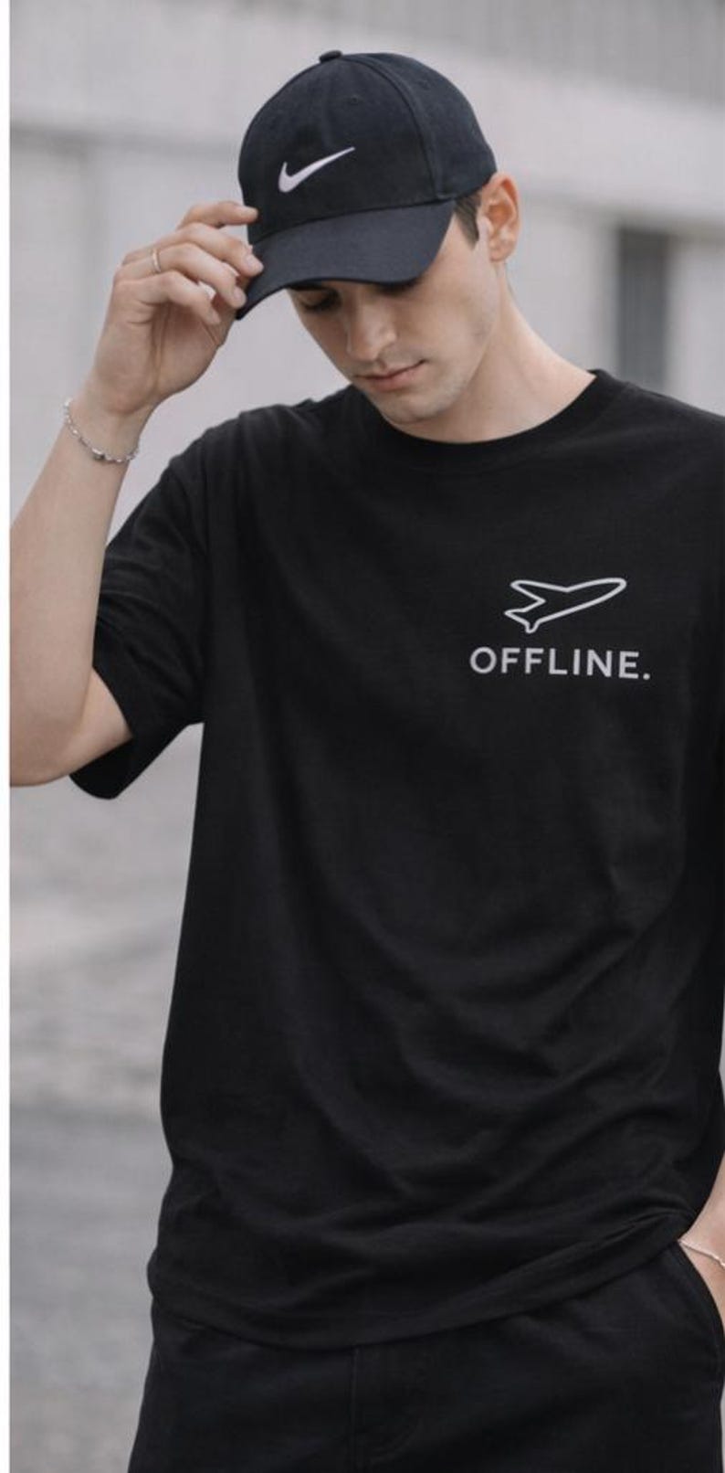 Puede incluir: Camiseta negra con un gr&aacute;fico de avi&oacute;n blanco y la palabra "OFFLINE". Se usa una gorra de b&eacute;isbol negra con el logotipo blanco de Nike. La imagen muestra a una persona usando la camiseta y la gorra.