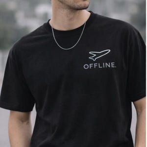 Puede incluir: Camiseta negra con un gr&aacute;fico de avi&oacute;n blanco y la palabra "OFFLINE". El modelo lleva una cadena y una pulsera de plata. Los pantalones son de color verde oliva. Estilo casual y moderno.