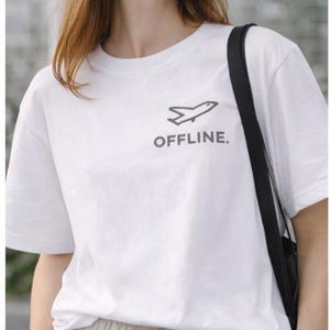 Puede incluir: Camiseta blanca con un peque&ntilde;o gr&aacute;fico de avi&oacute;n y la palabra "OFFLINE" en gris. La persona lleva pantalones beige con cord&oacute;n y un bolso negro. El fondo est&aacute; borroso.