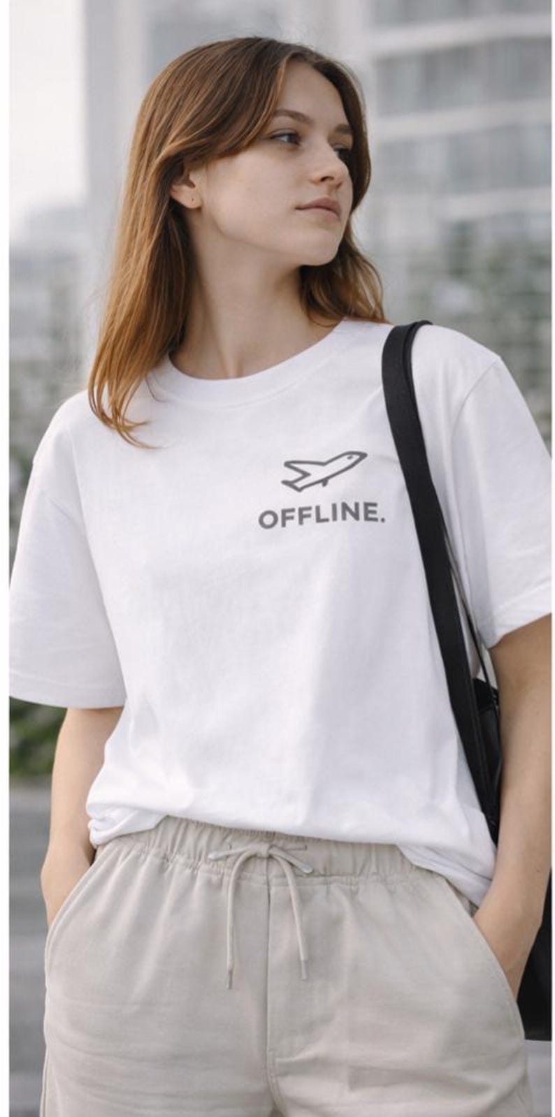 Puede incluir: Camiseta blanca con un peque&ntilde;o gr&aacute;fico de avi&oacute;n y la palabra "OFFLINE" en gris. La persona lleva pantalones beige con cord&oacute;n y un bolso negro. El fondo est&aacute; borroso.